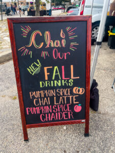 A hand drawn chalk sign saying "Chai Our New Fall Drinks" in swirling letters in orange, red and yellow. Below, drinks include a pumpkin spice chai latte and a pumpkin spice apple chaider.